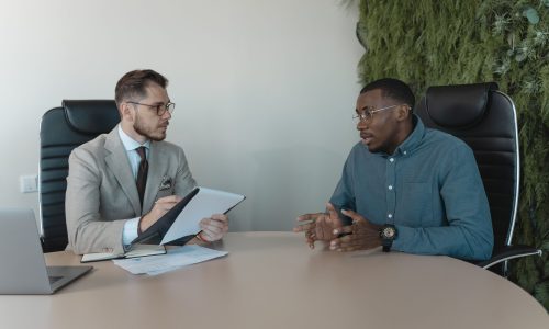 Two men engaged in a behavioral interview, seated at a table, discussing professional experiences and skills.
