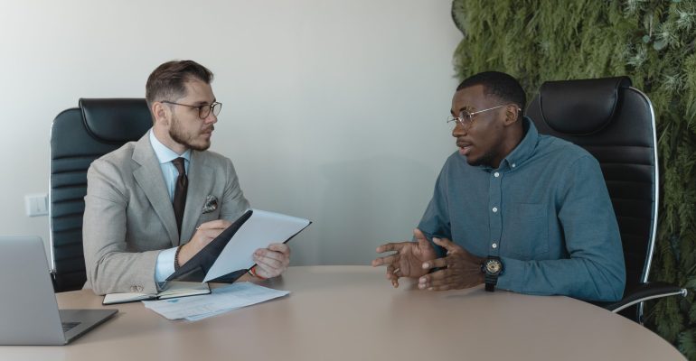 Two men engaged in a behavioral interview, seated at a table, discussing professional experiences and skills.